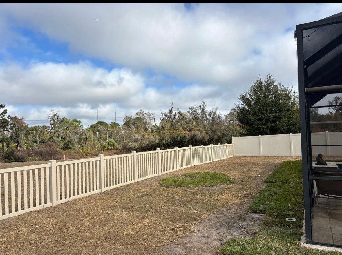 White corrugated metal fence running along a cleared dirt property with green trees and blue sky in background
